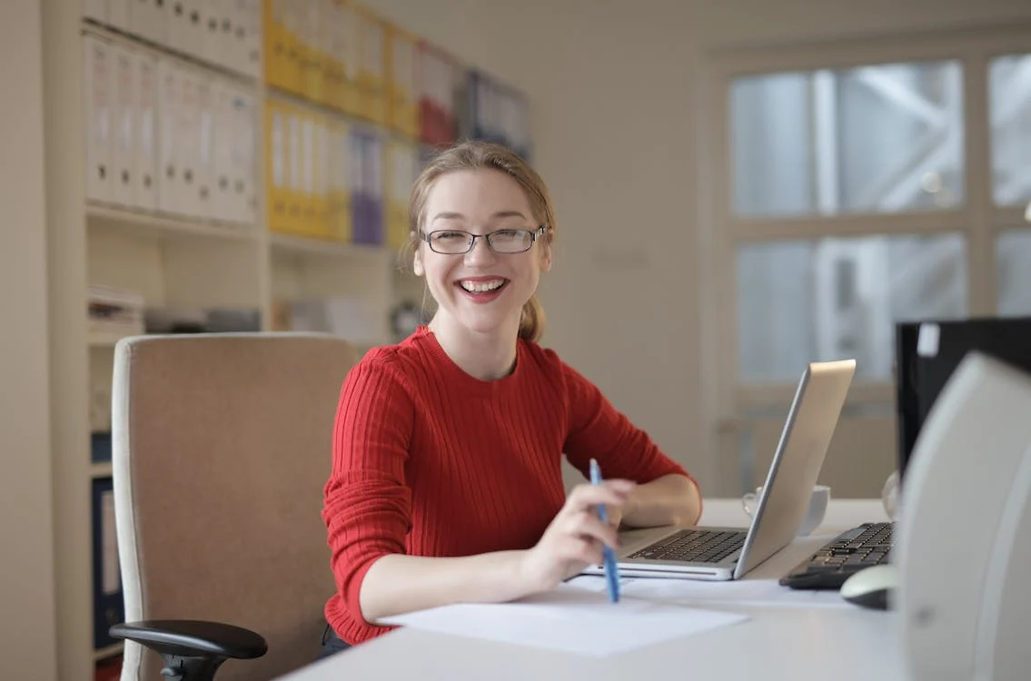Illustrative photo: a person at a desk in an office with files of papers on shelves behind, but despite the old school working setup seems infectiously positive and smiley